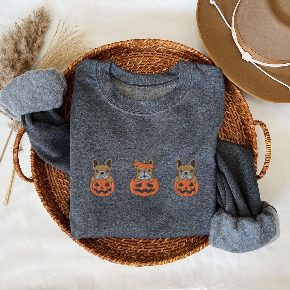 Frenchie Pumpkin Sweatshirt – Dark Heather Dark Heather crewneck sweatshirt folded in a wicker basket with a felt hat and dried grass, embroidered with three fawn French bulldogs peeking out of carved jack-o'-lanterns