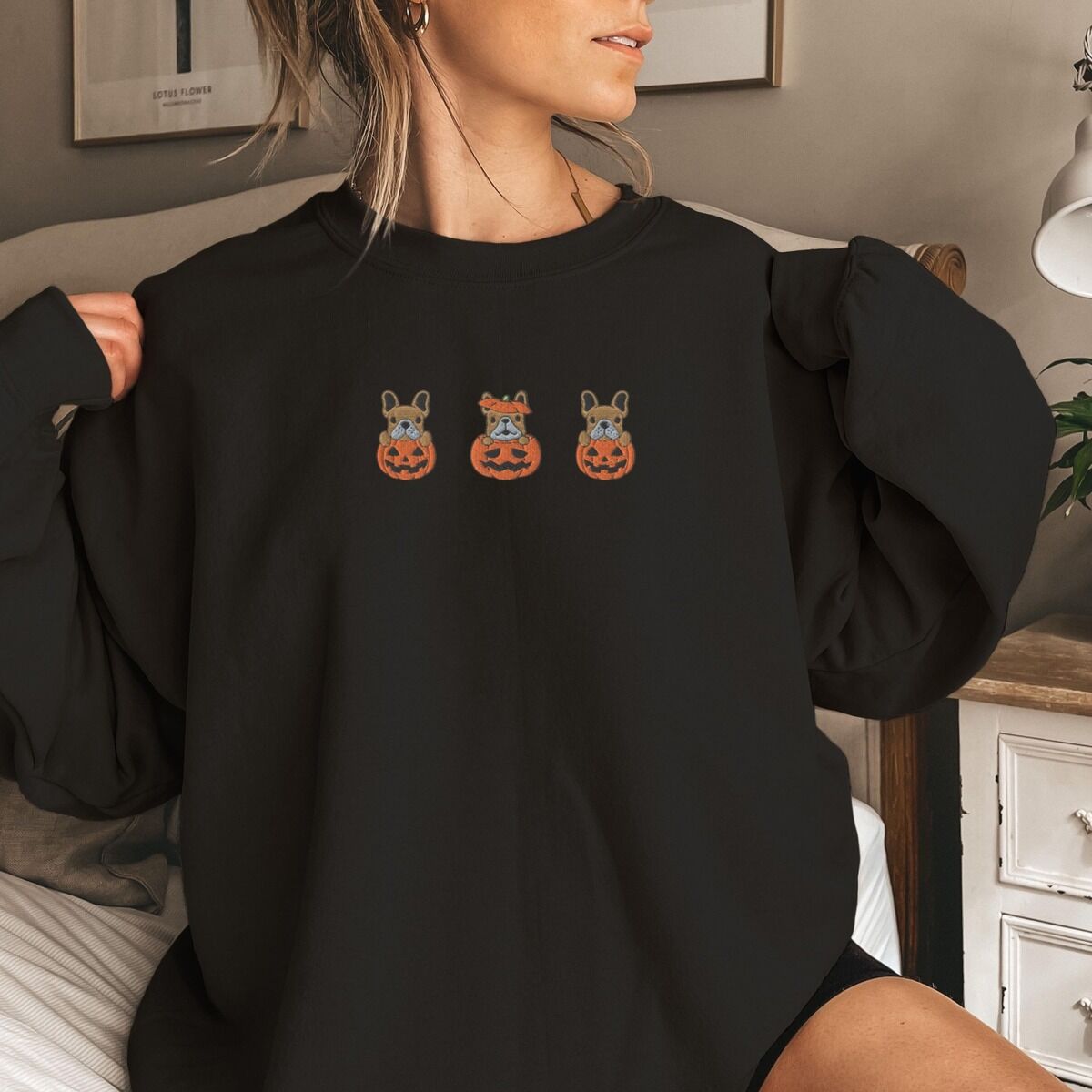 Frenchie Pumpkin Sweatshirt - worn Woman wearing a black crewneck sweatshirt in a bedroom, embroidered with three fawn French bulldogs peeking out of carved jack-o'-lanterns