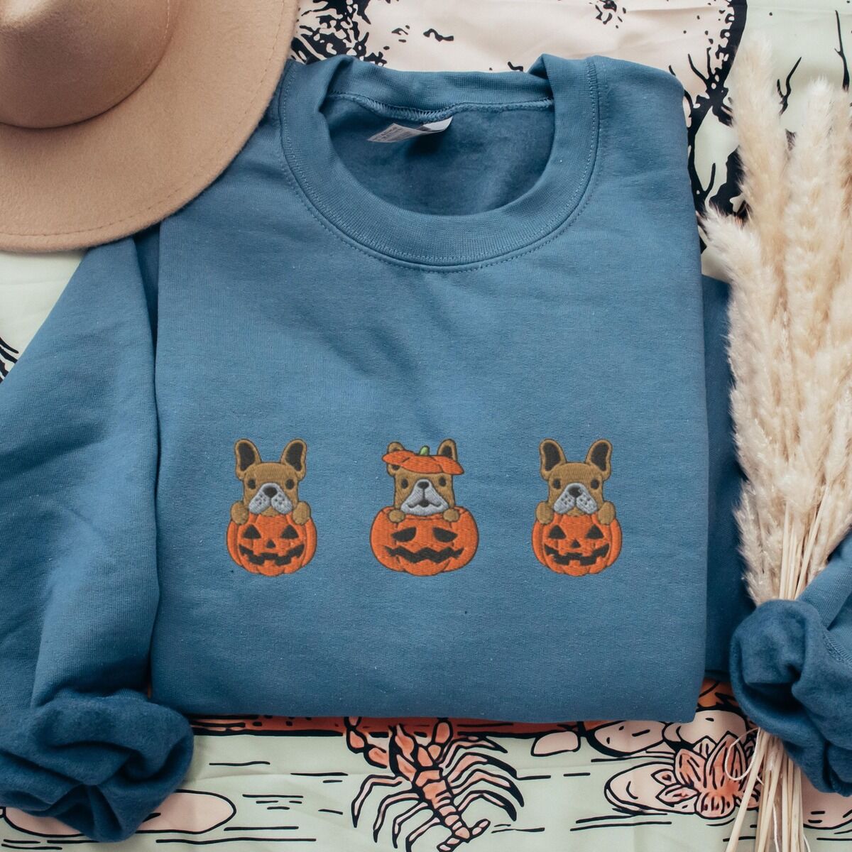 Frenchie Pumpkin Sweatshirt – Indigo Blue Indigo Blue crewneck sweatshirt laid flat on a printed backdrop with a felt hat and pampas grass, embroidered with three fawn French bulldogs peeking out of carved jack-o'-lanterns