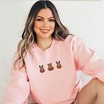 Smiling woman sitting in a Light Pink crewneck sweatshirt, embroidered with three fawn French bulldogs peeking out of carved jack-o'-lanterns