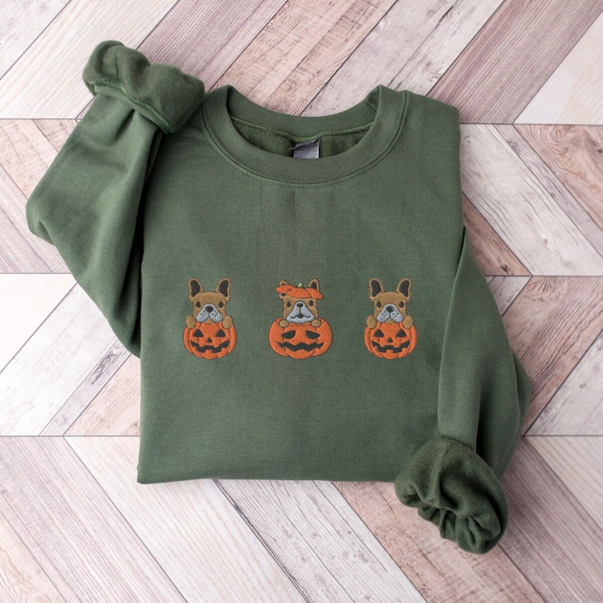 Frenchie Pumpkin Sweatshirt – Military Green Military Green crewneck sweatshirt laid flat on a chevron wood floor, embroidered with three fawn French bulldogs peeking out of carved jack-o'-lanterns