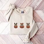 Sand crewneck sweatshirt laid flat on a chevron wood floor, embroidered with three fawn French bulldogs peeking out of carved jack-o'-lanterns