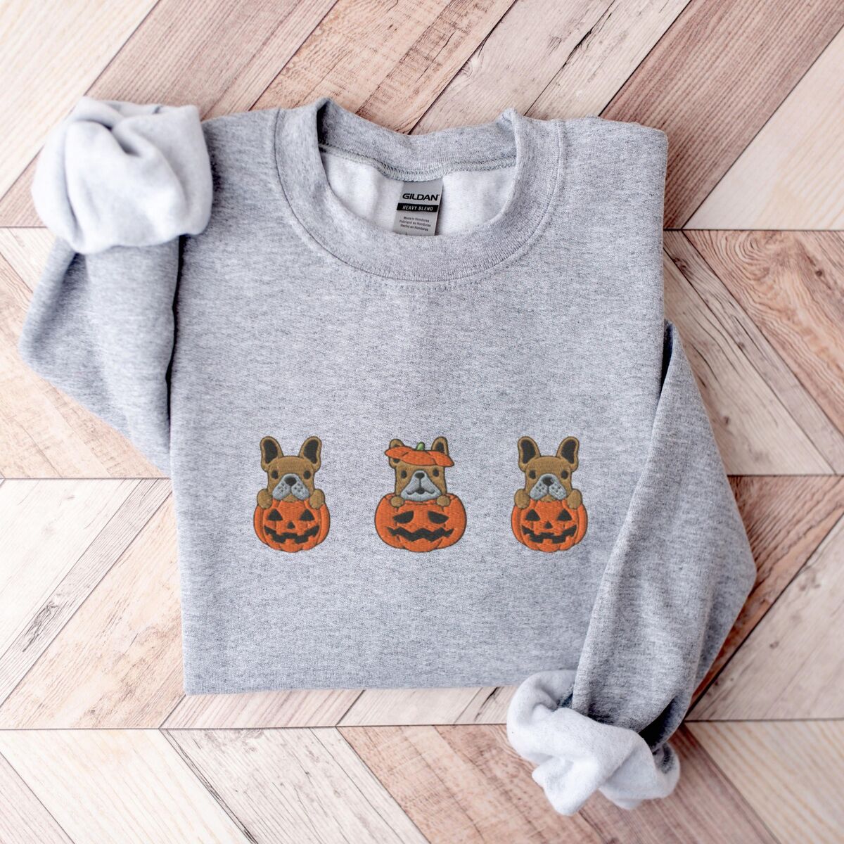 Frenchie Pumpkin Sweatshirt - Sport Grey Sport Grey crewneck sweatshirt laid flat on a chevron wood floor, embroidered with three fawn French bulldogs peeking out of carved jack-o'-lanterns