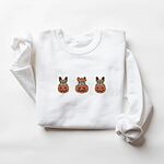 White crewneck sweatshirt laid flat on a white background, embroidered with three fawn French bulldogs peeking out of carved jack-o'-lanterns