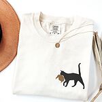 Ivory Comfort Colors T-shirt embroidered with a black cat walking off with a golden toupee in its mouth on the left chest