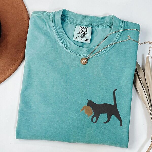 Seafoam Comfort Colors T-shirt embroidered with a black cat walking off with a golden toupee in its mouth on the left chest