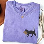 Violet Comfort Colors T-shirt embroidered with a black cat walking off with a golden toupee in its mouth on the left chest