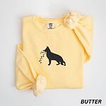 Butter crewneck sweatshirt laid flat with a rolled cuff, embroidered with a black German shepherd silhouette carrying a string of colored Christmas lights