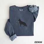 Denim crewneck sweatshirt laid flat with a rolled cuff, embroidered with a black German shepherd silhouette carrying a string of colored Christmas lights