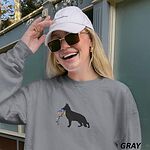 Laughing woman in a cap and sunglasses wearing a Grey crewneck sweatshirt embroidered with a black German shepherd silhouette carrying a string of colored Christmas lights