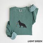 Light Green crewneck sweatshirt laid flat with a rolled cuff, embroidered with a black German shepherd silhouette carrying a string of colored Christmas lights