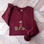 Maroon crewneck sweatshirt laid flat on a chevron wood floor, embroidered with a black and tan German shepherd in a Santa hat and red scarf beside a green polka dot gift box