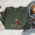 Military Green crewneck sweatshirt laid flat on a faux fur rug with pinecones and a knit blanket, embroidered with a black and tan German shepherd in a Santa hat and red scarf beside a green polka dot gift box