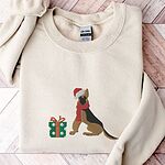 Sand crewneck sweatshirt laid flat on a pale wood floor, embroidered with a black and tan German shepherd in a Santa hat and red scarf beside a green polka dot gift box