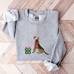 Sport Grey crewneck sweatshirt laid flat on a chevron wood floor, embroidered with a black and tan German shepherd in a Santa hat and red scarf beside a green polka dot gift box