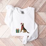 White crewneck sweatshirt laid flat on a chevron wood floor, embroidered with a black and tan German shepherd in a Santa hat and red scarf beside a green polka dot gift box