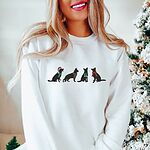 Woman beside a snowy Christmas tree in a White crewneck sweatshirt embroidered with four black German shepherd silhouettes in a Santa hat, striped scarves and a red bow
