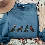 Indigo Blue crewneck sweatshirt laid flat with dried pampas grass and a hat, embroidered with four black German shepherd silhouettes in a Santa hat, striped scarves and a red bow