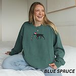 Woman smiling on a bed in a Blue Spruce crewneck sweatshirt embroidered with a German shorthaired pointer in a Santa hat holding Christmas lights