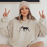 Woman in a beanie making peace signs in a Sand crewneck sweatshirt embroidered with a black German shorthaired pointer in a Santa hat carrying a string of Christmas lights