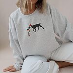 Woman sitting in a grey crewneck sweatshirt embroidered with a black German shorthaired pointer in a Santa hat carrying a string of Christmas lights