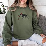 Woman on a sofa in a Military Green crewneck sweatshirt embroidered with a black German shorthaired pointer in a Santa hat carrying a string of Christmas lights