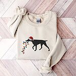 Sand crewneck sweatshirt laid flat on wood, embroidered with a black German shorthaired pointer in a Santa hat carrying a string of Christmas lights
