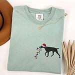 Bay T-shirt laid flat with a necklace and a straw hat, embroidered with a black German shorthaired pointer in a Santa hat carrying a string of Christmas lights on the left chest