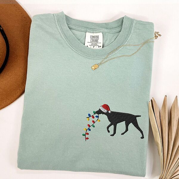 Bay T-shirt laid flat with a necklace and a straw hat, embroidered with a black German shorthaired pointer in a Santa hat carrying a string of Christmas lights on the left chest