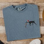 Blue Jean T-shirt laid flat on wood beside white sneakers, embroidered with a black German shorthaired pointer in a Santa hat carrying a string of Christmas lights on the left chest