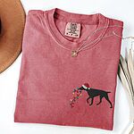Crimson T-shirt laid flat with a necklace, embroidered with a black German shorthaired pointer in a Santa hat carrying a string of Christmas lights on the left chest