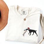 Ivory T-shirt laid flat with a necklace, embroidered with a black German shorthaired pointer in a Santa hat carrying a string of Christmas lights on the left chest