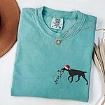 Seafoam T-shirt laid flat with a necklace, embroidered with a black German shorthaired pointer in a Santa hat carrying a string of Christmas lights on the left chest