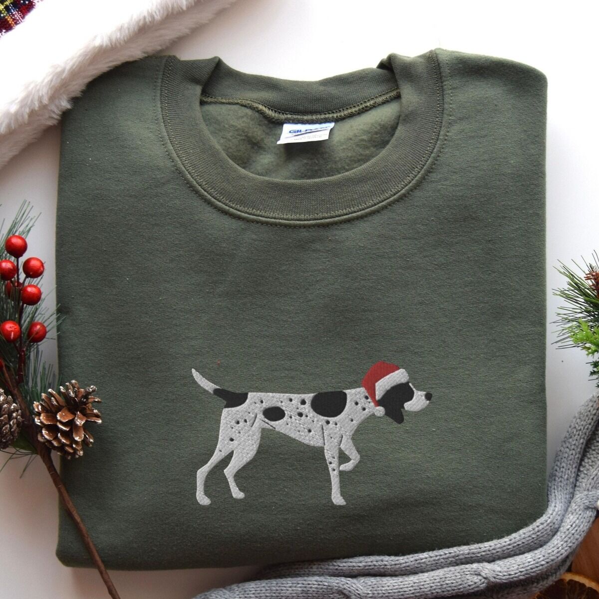 German Shorthaired Pointer Christmas Sweatshirt – Military Green Military Green crewneck sweatshirt laid flat with pine cones, red berries and a grey knit blanket, embroidered with a black and white ticked German shorthaired pointer in a red Santa hat