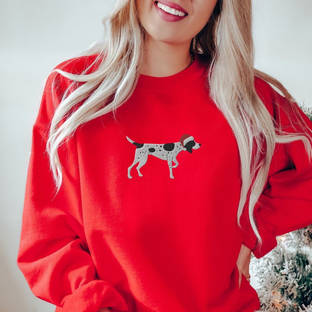 German Shorthaired Pointer Christmas Sweatshirt - Red Woman by a Christmas tree in a Red crewneck sweatshirt embroidered with a black and white ticked German shorthaired pointer in a red Santa hat