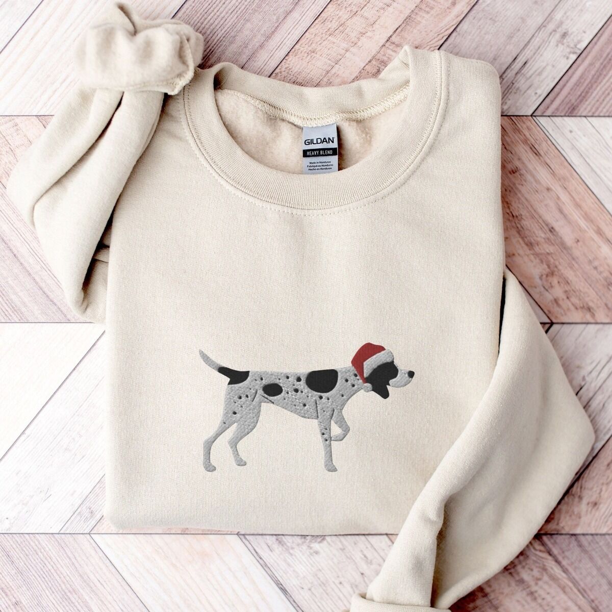 German Shorthaired Pointer Christmas Sweatshirt – Sand Sand crewneck sweatshirt laid flat on a herringbone wood floor, embroidered with a black and white ticked German shorthaired pointer in a red Santa hat