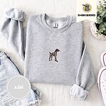 Ash crewneck sweatshirt folded with denim shorts, white sneakers and a potted plant, embroidered with a standing liver and white German shorthaired pointer in a tan collar