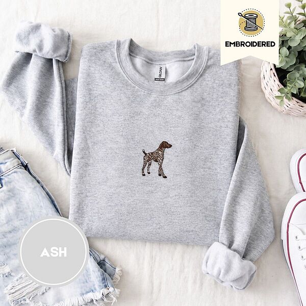 Ash crewneck sweatshirt folded with denim shorts, white sneakers and a potted plant, embroidered with a standing liver and white German shorthaired pointer in a tan collar