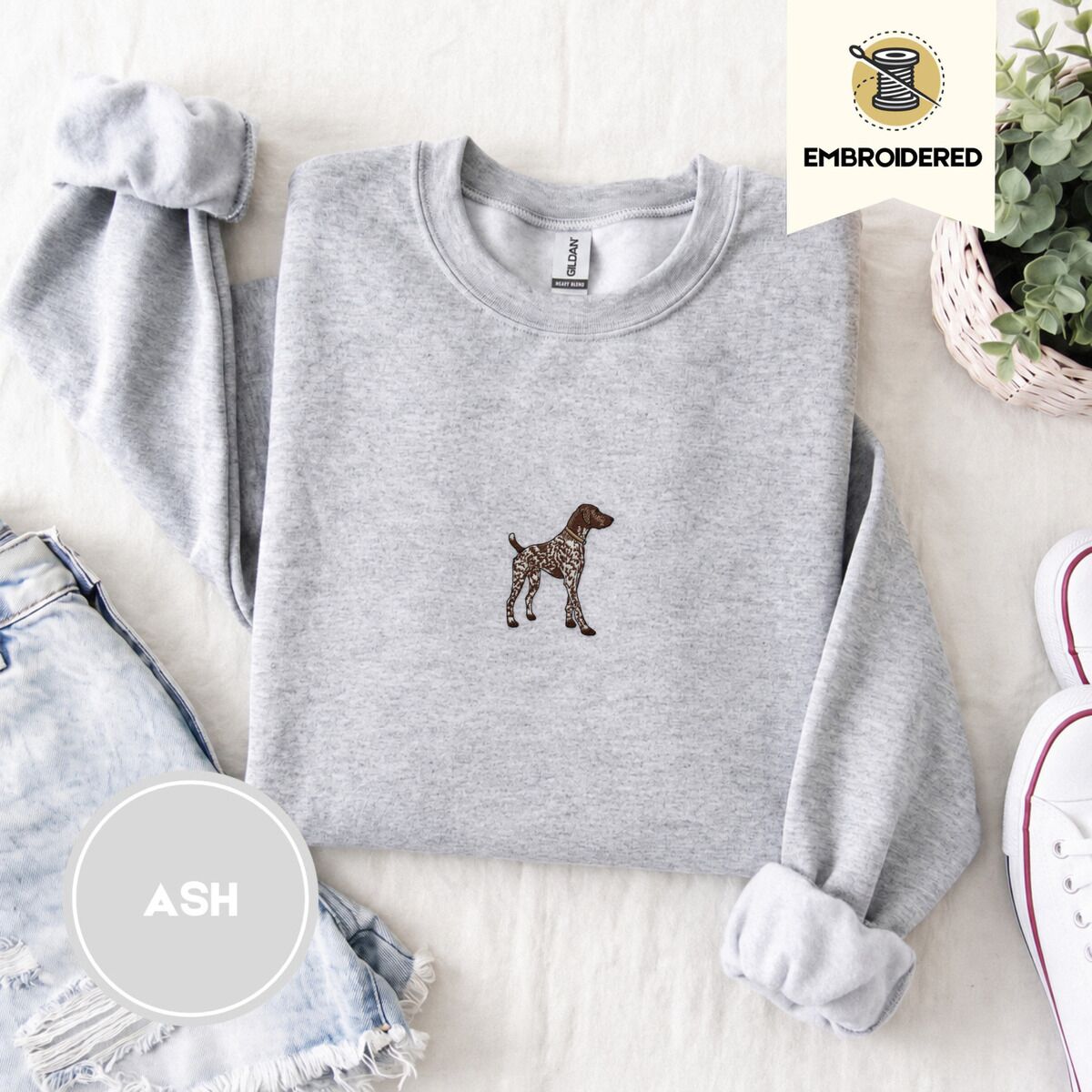 German Shorthaired Pointer Sweatshirt – Ash Ash crewneck sweatshirt folded with denim shorts, white sneakers and a potted plant, embroidered with a standing liver and white German shorthaired pointer in a tan collar