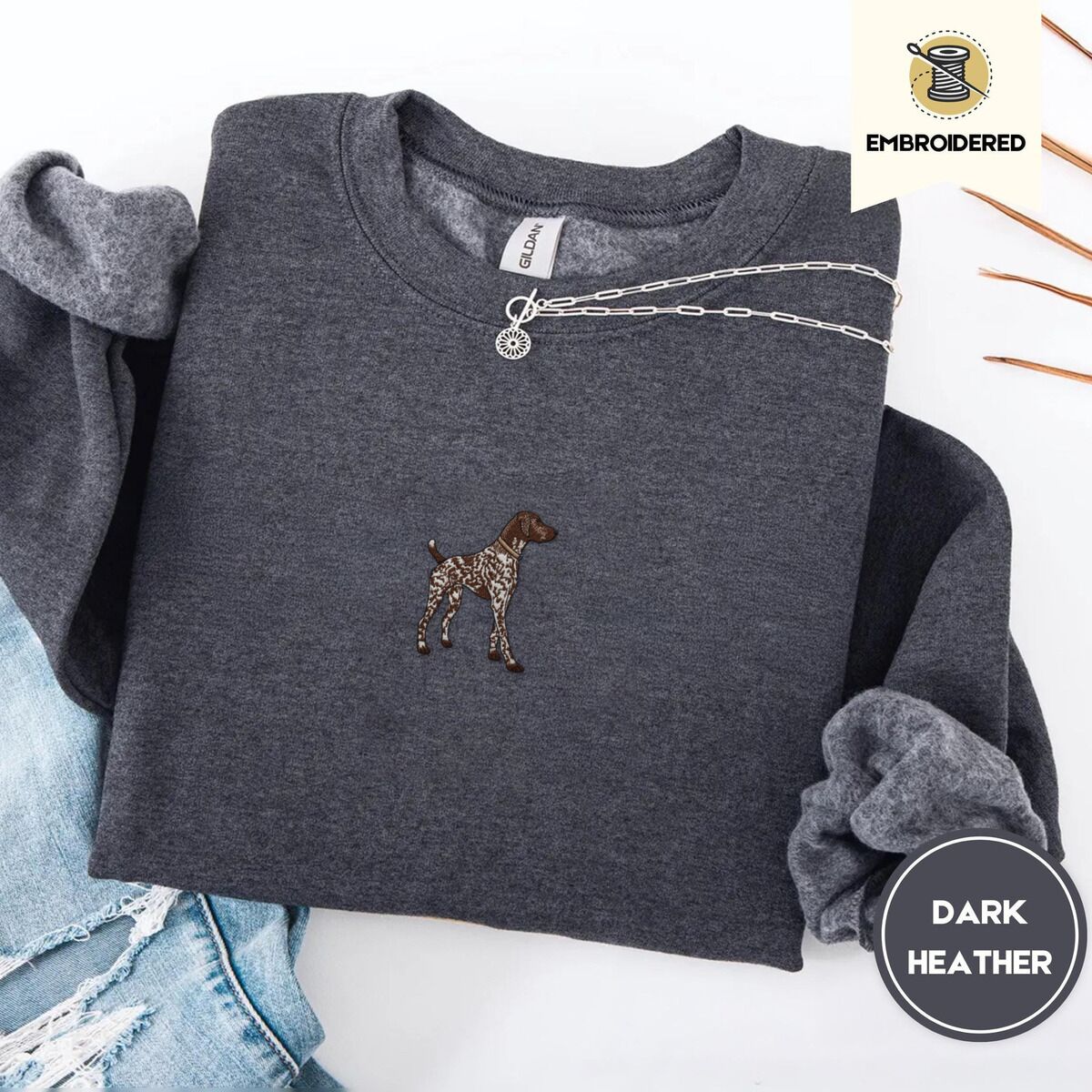 German Shorthaired Pointer Sweatshirt – Dark Heather Dark Heather crewneck sweatshirt folded with ripped jeans and a silver chain necklace, embroidered with a standing liver and white German shorthaired pointer in a tan collar