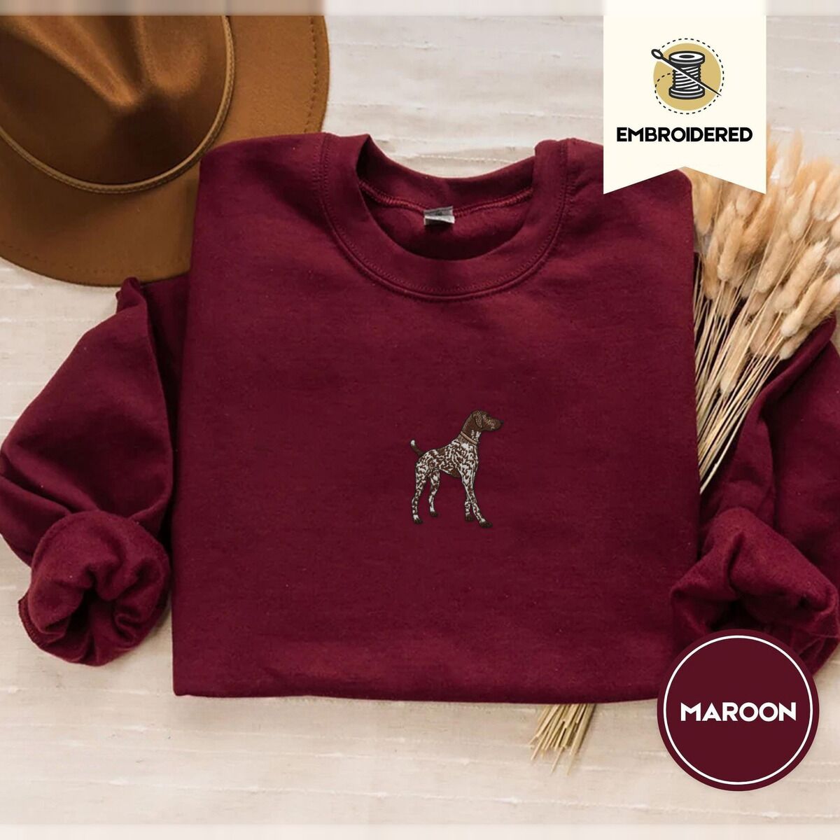 German Shorthaired Pointer Sweatshirt – Maroon Maroon crewneck sweatshirt with a brown felt hat and dried bunny tail grass, embroidered with a standing liver and white German shorthaired pointer in a tan collar