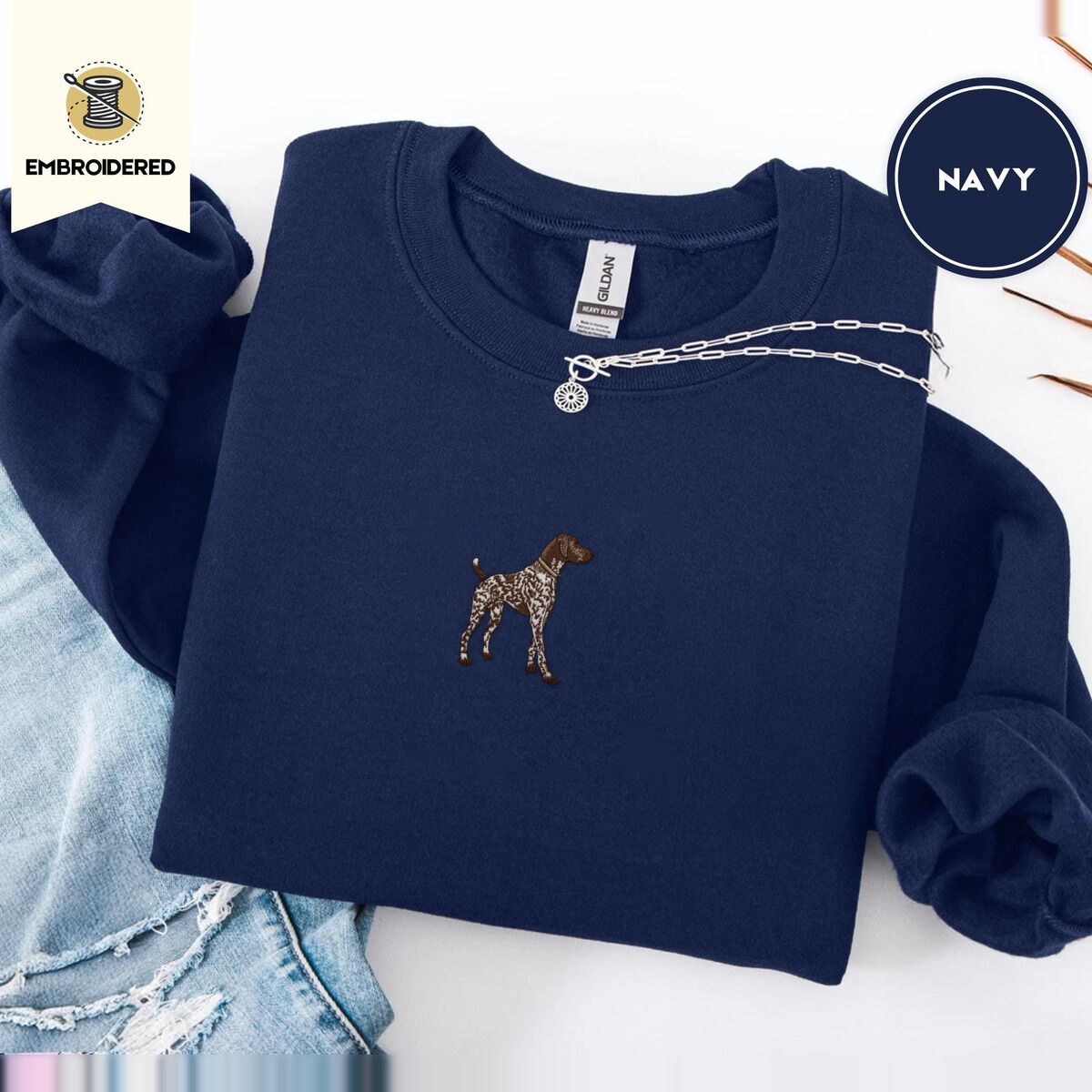 German Shorthaired Pointer Sweatshirt – Navy Navy crewneck sweatshirt folded with ripped jeans and a silver chain necklace, embroidered with a standing liver and white German shorthaired pointer in a tan collar