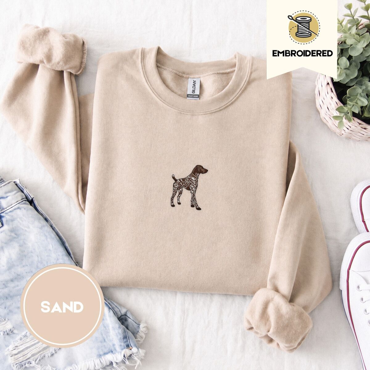 German Shorthaired Pointer Sweatshirt – Sand Sand crewneck sweatshirt folded with denim shorts, white sneakers and a potted plant, embroidered with a standing liver and white German shorthaired pointer in a tan collar