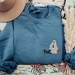 Indigo Blue crewneck sweatshirt laid flat on a patterned throw with a felt hat and pampas grass, embroidered with a ghost sipping iced coffee with a black pit bull in a play bow on the left chest