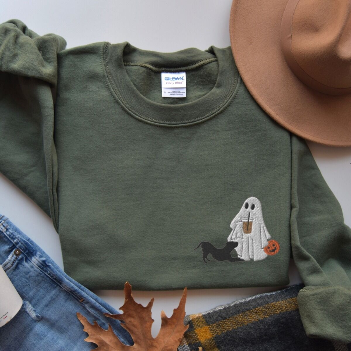 Ghost and Pitbull Sweatshirt – Military Green Military Green crewneck sweatshirt laid flat with jeans, fall leaves, a plaid scarf and a felt hat, embroidered with a ghost sipping iced coffee with a black pit bull in a play bow on the left chest