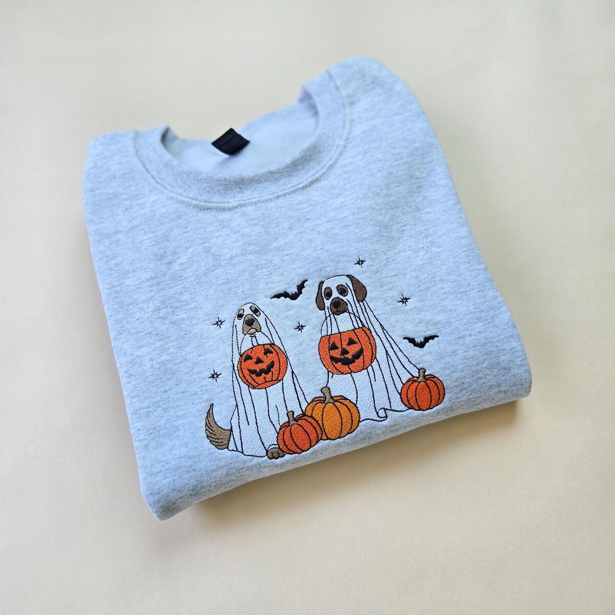 Ghost Dogs with Pumpkins Sweatshirt – Ash Ash crewneck sweatshirt folded on a pale yellow background, embroidered with two dogs in white ghost sheets holding jack-o'-lantern buckets among pumpkins, with bats and stars