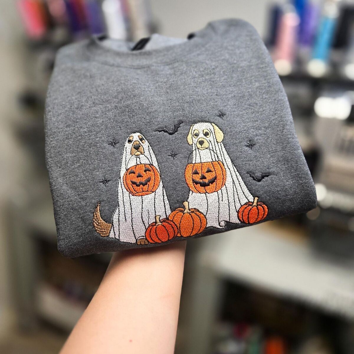 Ghost Dogs with Pumpkins Sweatshirt – Custom Coat Colors on Dark Heather Dark Heather ghost dogs with pumpkins sweatshirt held up by hand, stitched with custom coat colors: a golden dog and a yellow dog in ghost sheets