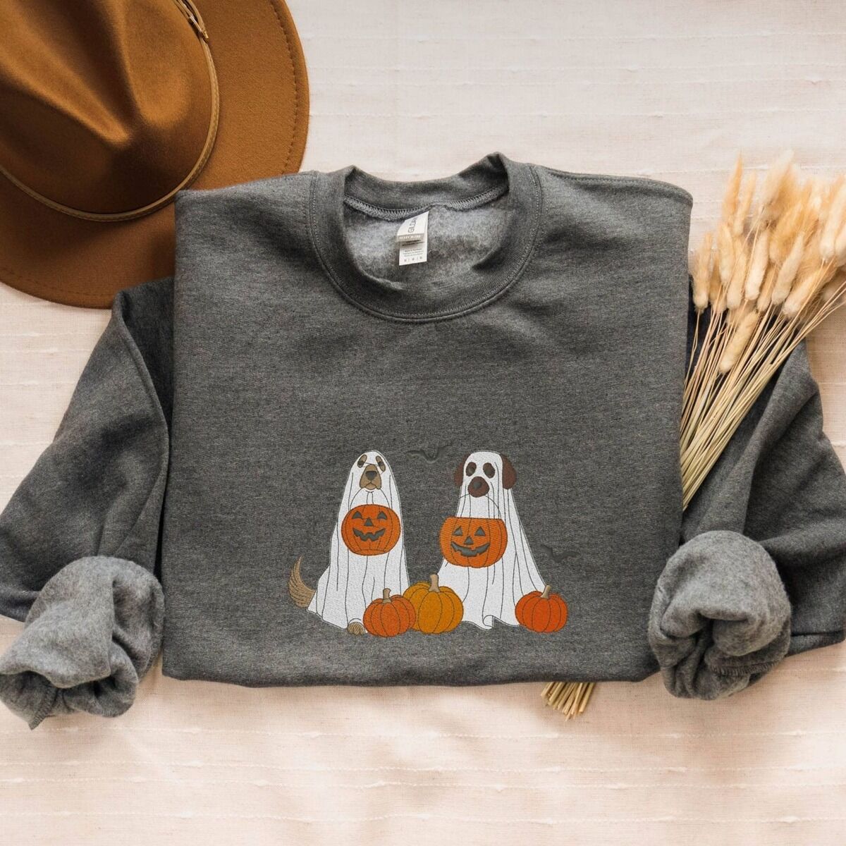 Ghost Dogs with Pumpkins Sweatshirt – Dark Heather Dark Heather crewneck sweatshirt folded with a brown felt hat and dried bunny tail grass, embroidered with two dogs in white ghost sheets holding jack-o'-lantern buckets among pumpkins, with bats and stars