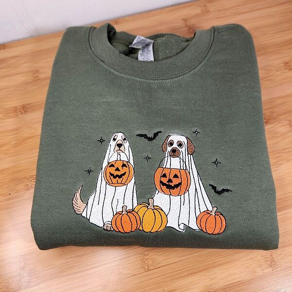 Military Green crewneck sweatshirt folded on a wooden table, embroidered with two dogs in white ghost sheets holding jack-o'-lantern buckets among pumpkins, with bats and stars