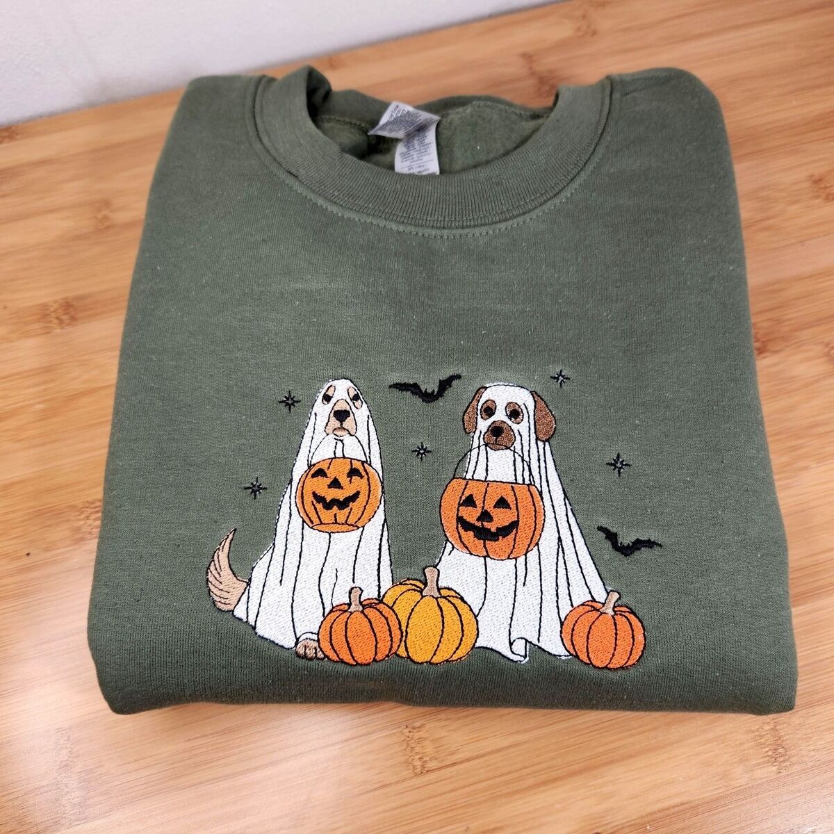 Ghost Dogs with Pumpkins Sweatshirt – Military Green Military Green crewneck sweatshirt folded on a wooden table, embroidered with two dogs in white ghost sheets holding jack-o'-lantern buckets among pumpkins, with bats and stars